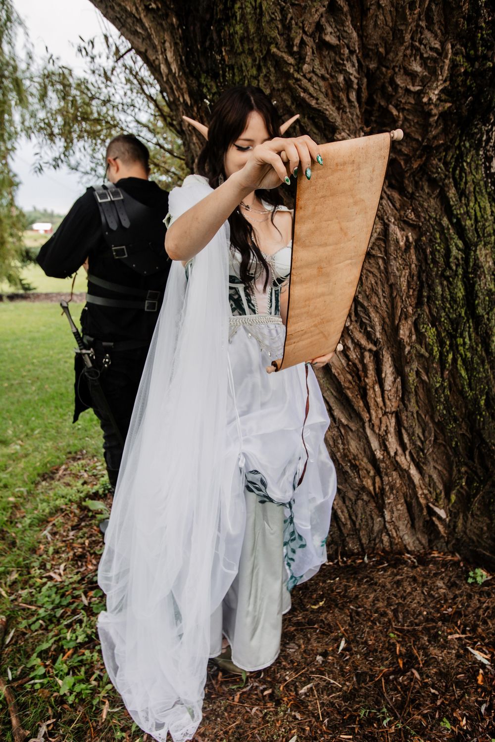 A couple stands at a tree while one reads from a scroll, wearing contrasting black and white outfits.