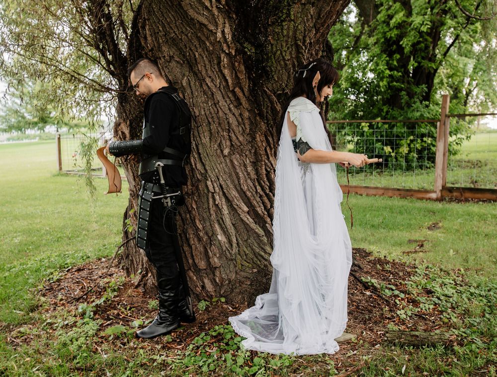 A couple stands on opposite sides of a large tree trunk in contrasting black and white wedding attire.