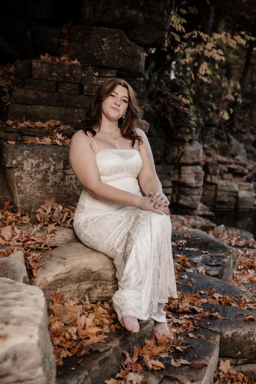 Romantic senior  portrait in white dress among autumn leaves and stone walls.