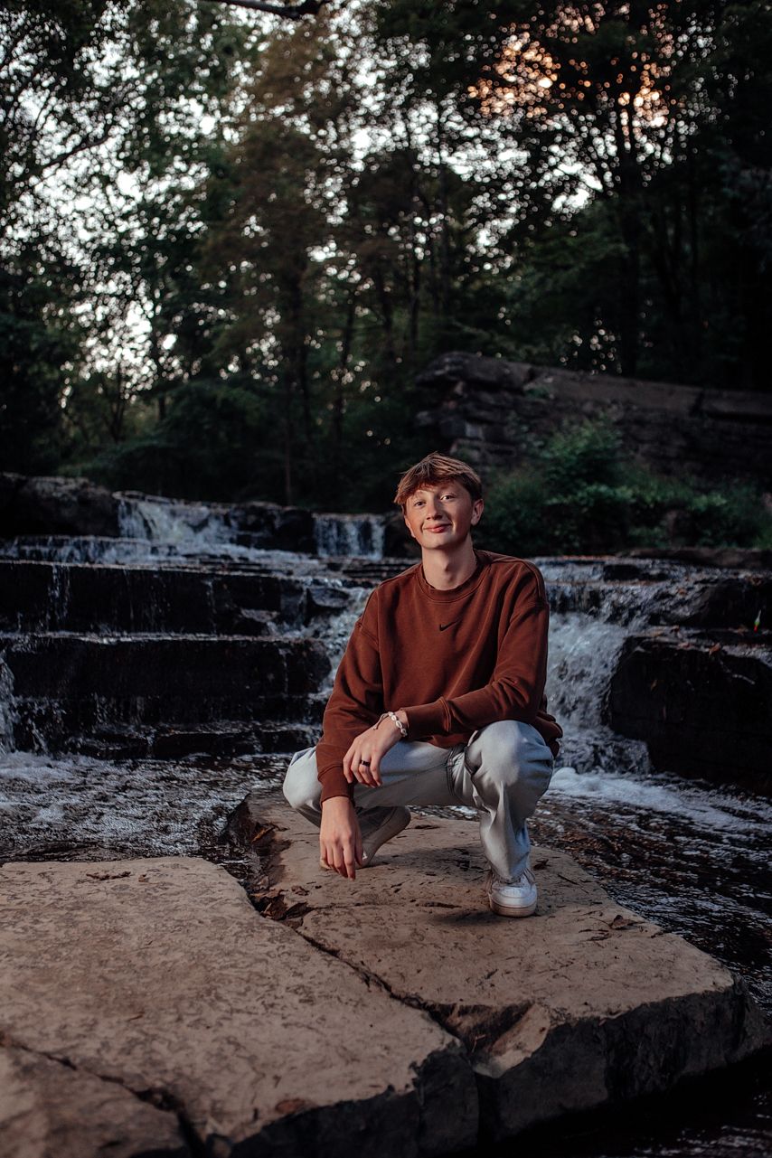 Relaxed senior outdoor portrait by waterfall rocks at sunset wearing brown sweater.