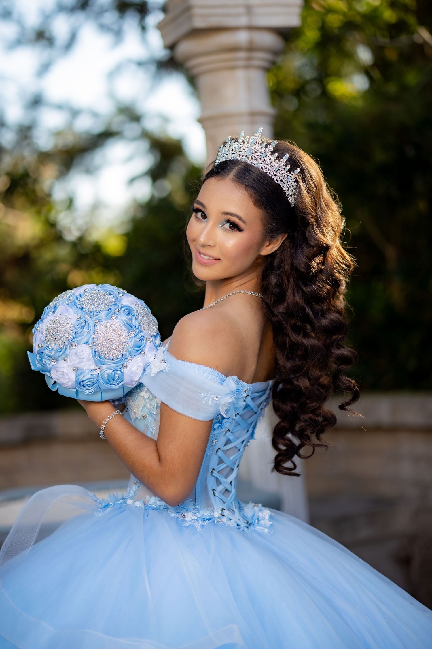 Young woman in a blue off-shoulder quinceañera ball gown with tiara holding a blue rose bouquet outdoors.