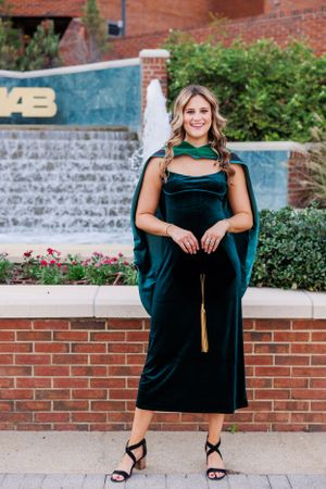 A graduate poses in an elegant dark green velvet graduation gown in front of a campus fountain.