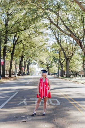 Graduate in red cap and gown poses on tree-lined campus street.