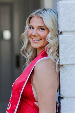 Portrait against white brick wall featuring graduate in red academic regalia at University of Alabama