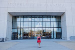 Modern glass entrance to University of Alabama building with graduate posing in red dress.