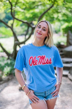 A person in an Ole Miss t-shirt and denim shorts poses outdoors on a sunny day.