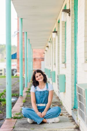 Person in denim overalls sits cross-legged on porch with turquoise pillars.