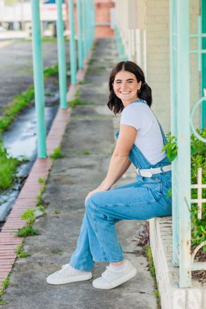 Person in denim overalls sits on ledge with turquoise columns in background.