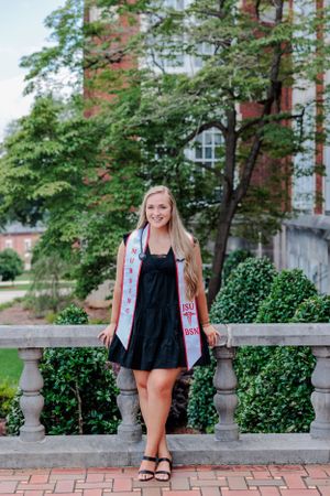 Graduate in a black dress and graduation stole poses on campus grounds with trees and brick building behind.