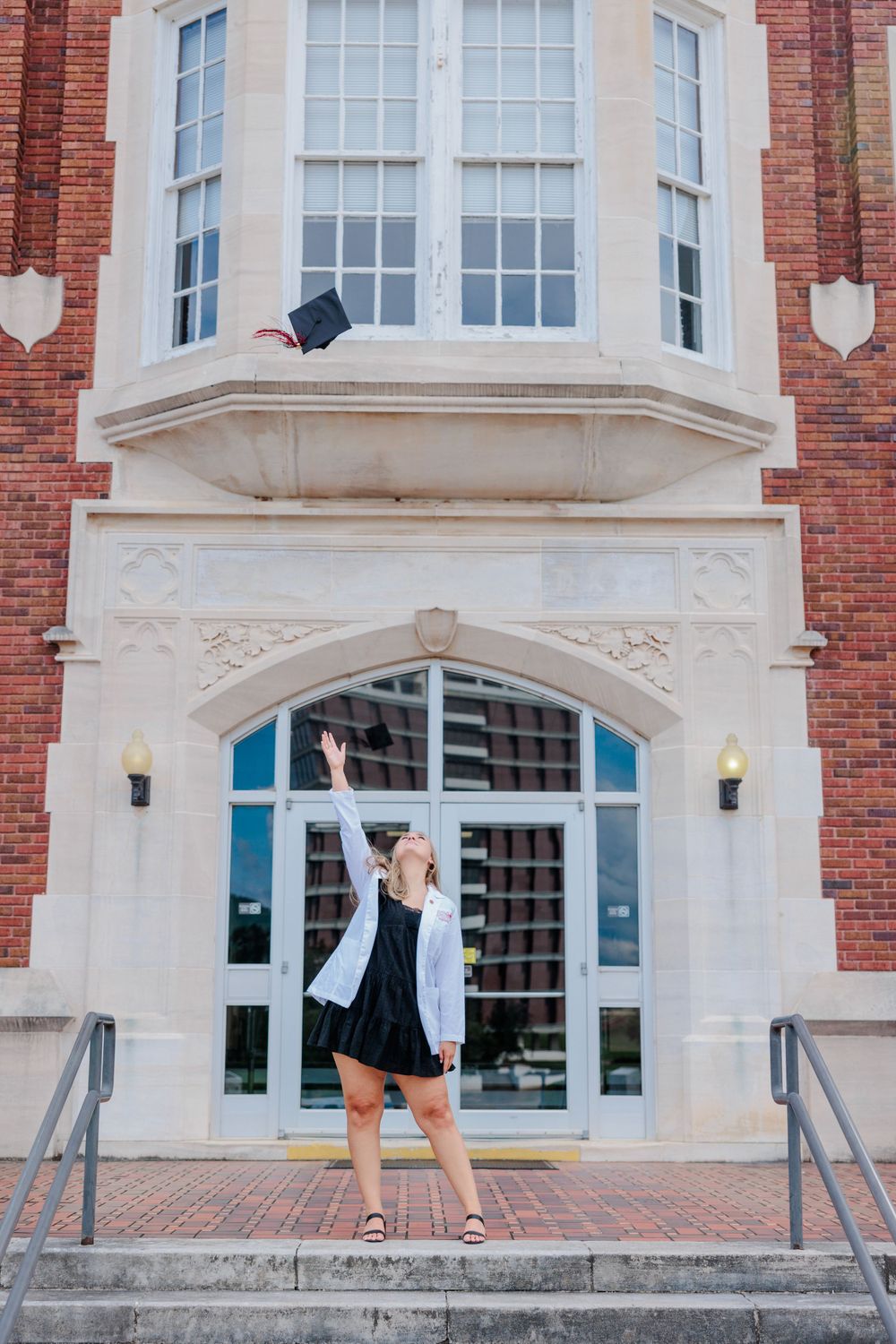 Graduate throws cap in front of ornate brick building entrance with arched doorway and windows.