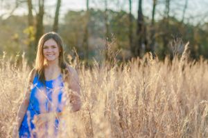 Person in blue dress stands in field of tall golden grass during sunset.