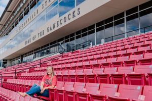 Graduate poses in stadium seats wearing casual attire against blue sky backdrop at JSU.