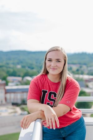Student in red university shirt leans on railing with scenic mountain view in background at JSU.