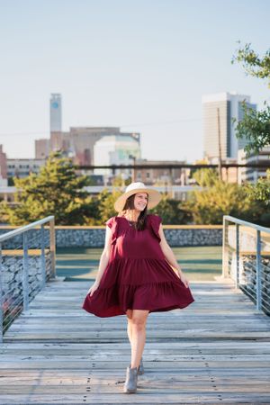 Person in flowing burgundy dress and hat twirling on wooden boardwalk with cityscape behind.