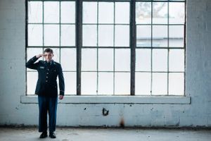 Figure in navy uniform stands by industrial windows in empty room.