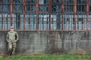 Military personnel in camouflage uniform stands against industrial concrete wall with metal bars.