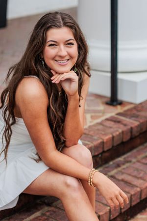 A portrait of someone in a white dress sitting on brick steps with a warm smile.