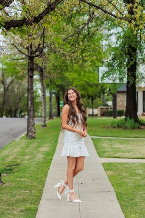A person in a white dress poses on a sidewalk lined with spring trees.