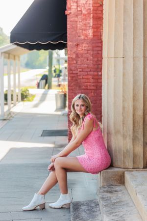 A person in a pink dress sits on stone steps near a brick building with black awning.