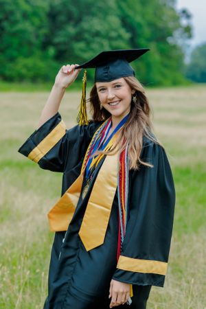 Graduate in cap and gown with gold stole and honor cords poses in grassy field.