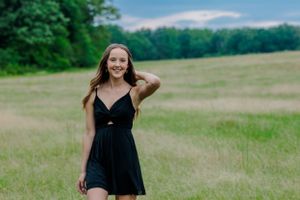 A woman in a black summer dress poses in a sunny meadow with trees in the background.