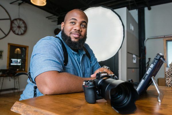 A photographer with a beard wearing a blue shirt sits at a wooden table with professional camera equipment and lighting.
