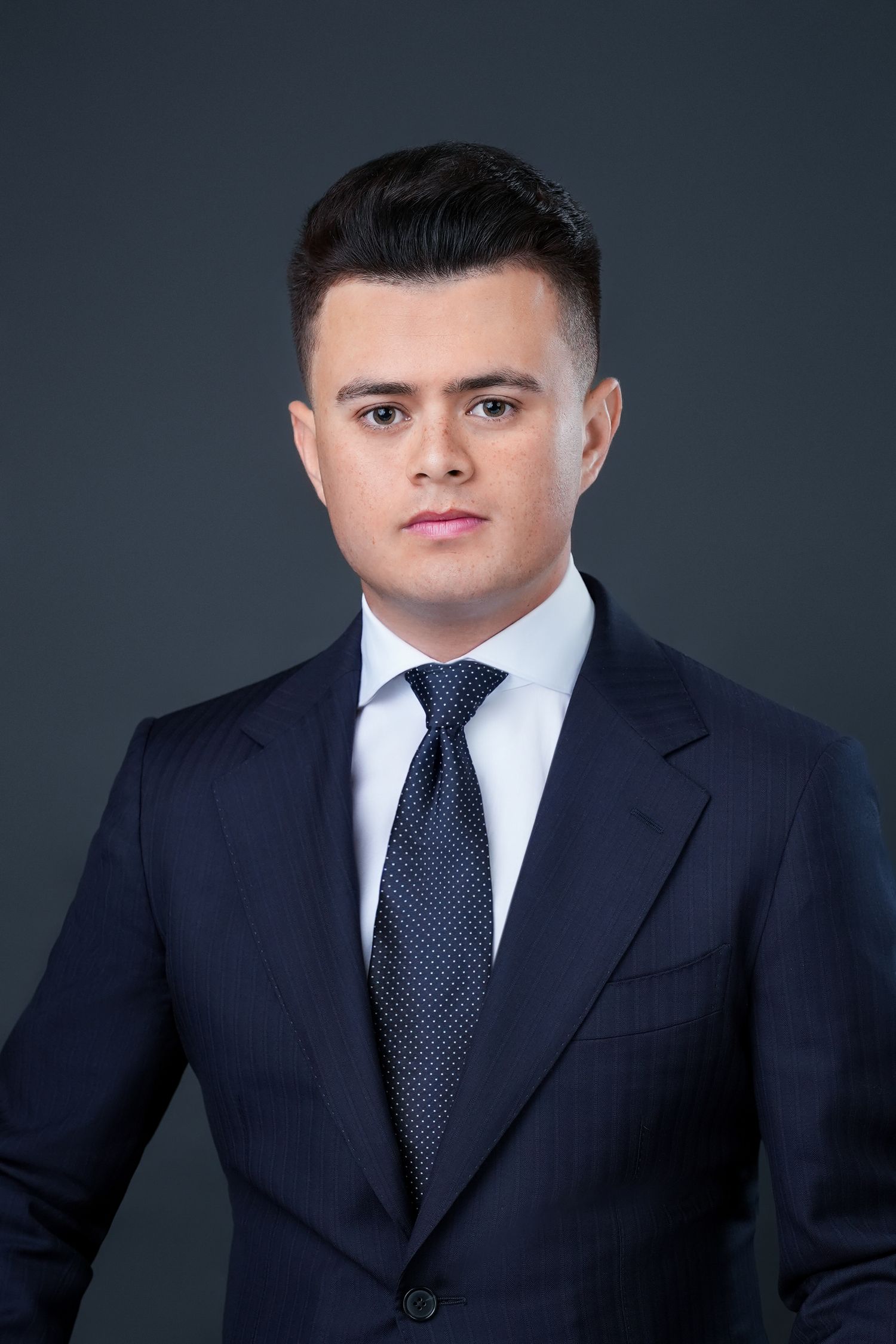 Professional headshot of person in navy suit and polka dot tie against dark background.