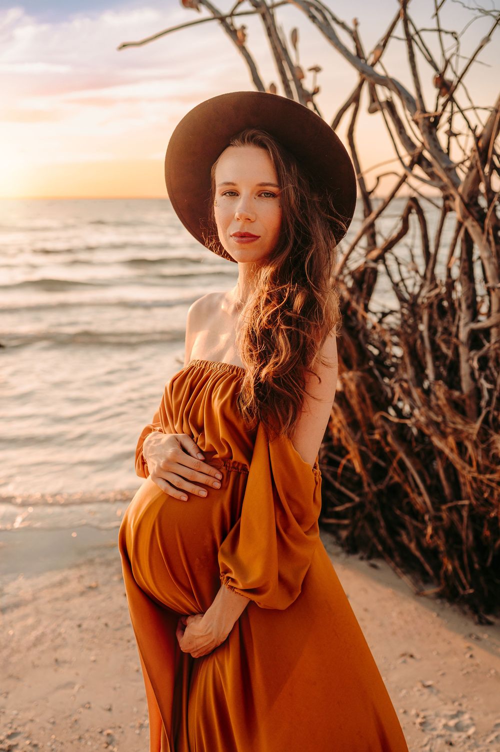 Woman in an orange, flowing dress standing on the beach at sunset, in a brown hat, holding her baby bump with both hands.