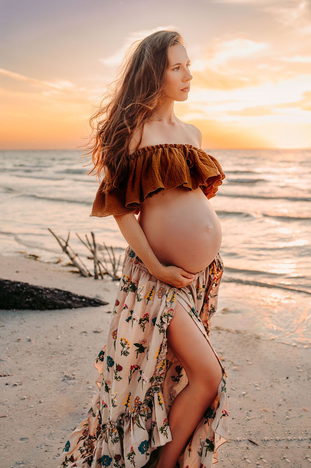 Backlit maternity photo of a woman in a soft two piece gown looking out to the ocean, cradling her belly.