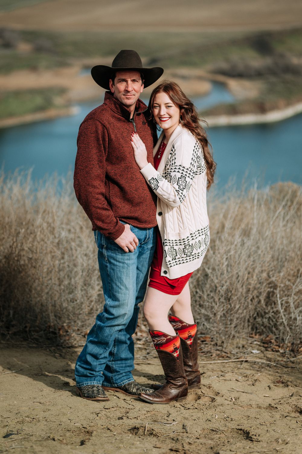 Couple posing outdoors in western attire, man in cowboy hat and woman in white cardigan with boots, scenic river backdrop.