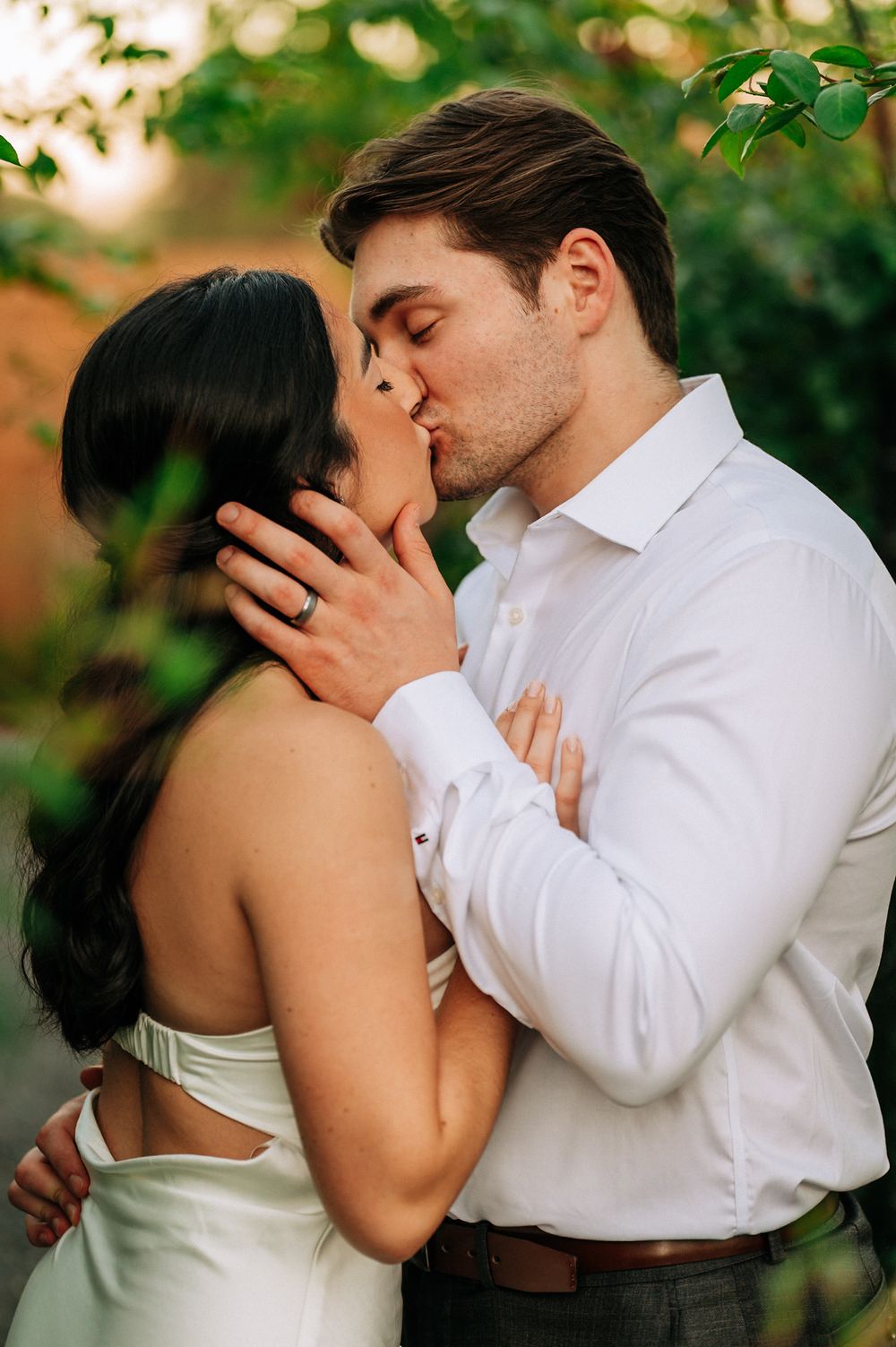 A couple shares a romantic kiss outdoors, the woman wearing a white dress with an engagement ring visible on her hand.