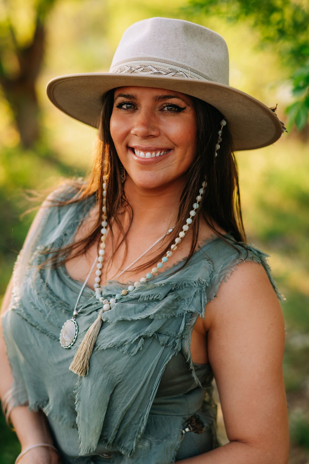 A person wearing a grey wide-brimmed hat and sage green dress smiles outdoors with boho style jewelry and natural lighting.