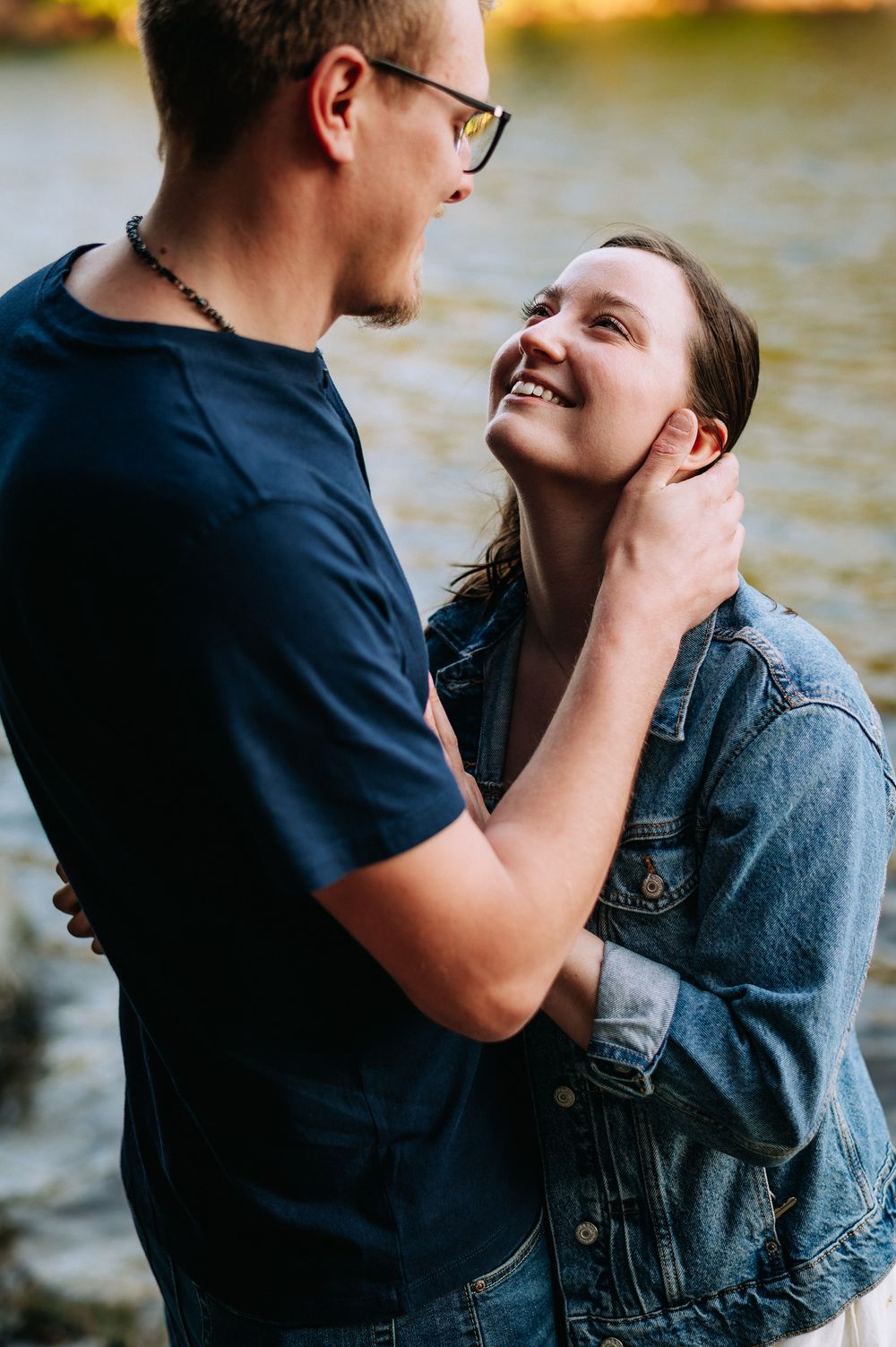 Romantic couple embraces by a lake during sunset, sharing an intimate moment while wearing casual denim and navy outfits.