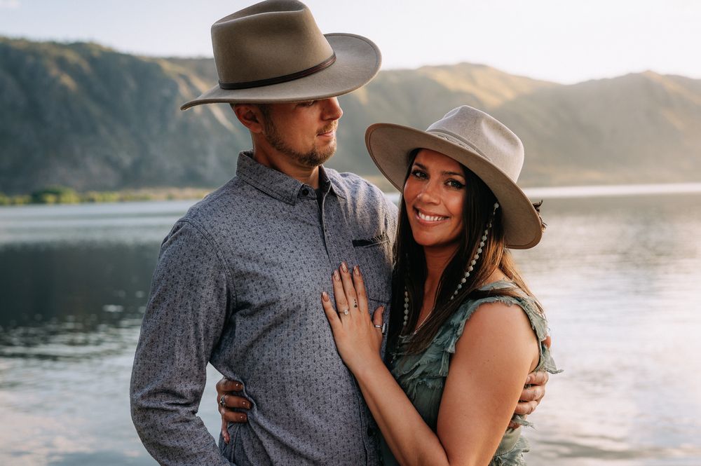 Happy couple in cowboy hats embrace by scenic lake at sunset with mountains in background.