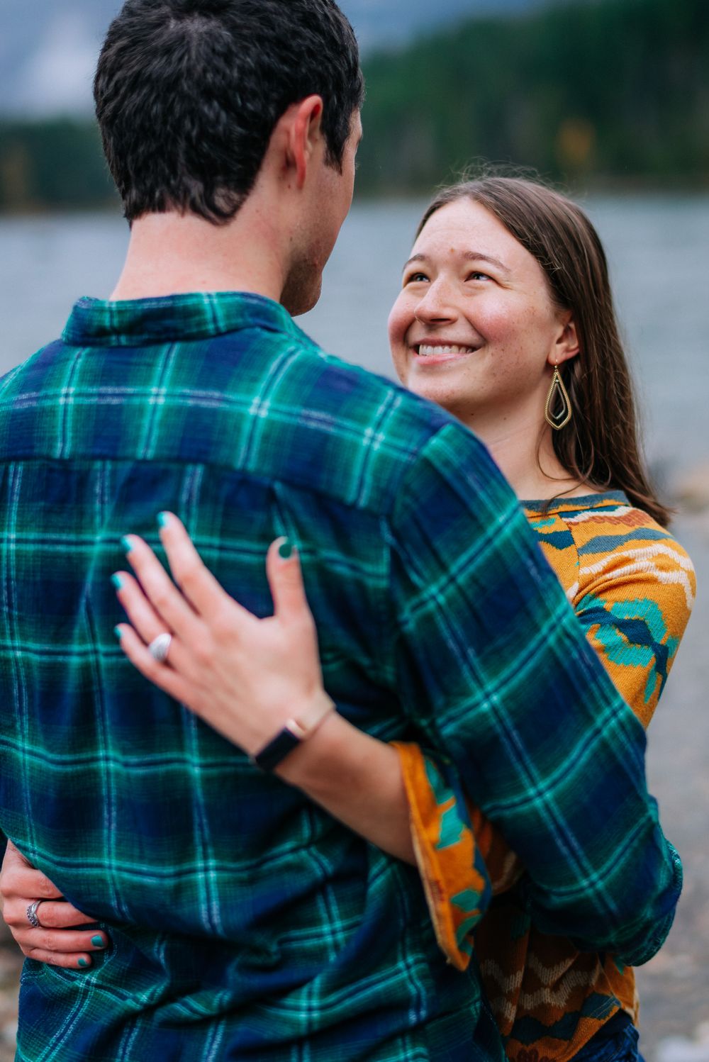 A couple embraces outdoors while wearing plaid and yellow, sharing a joyful moment by the water.