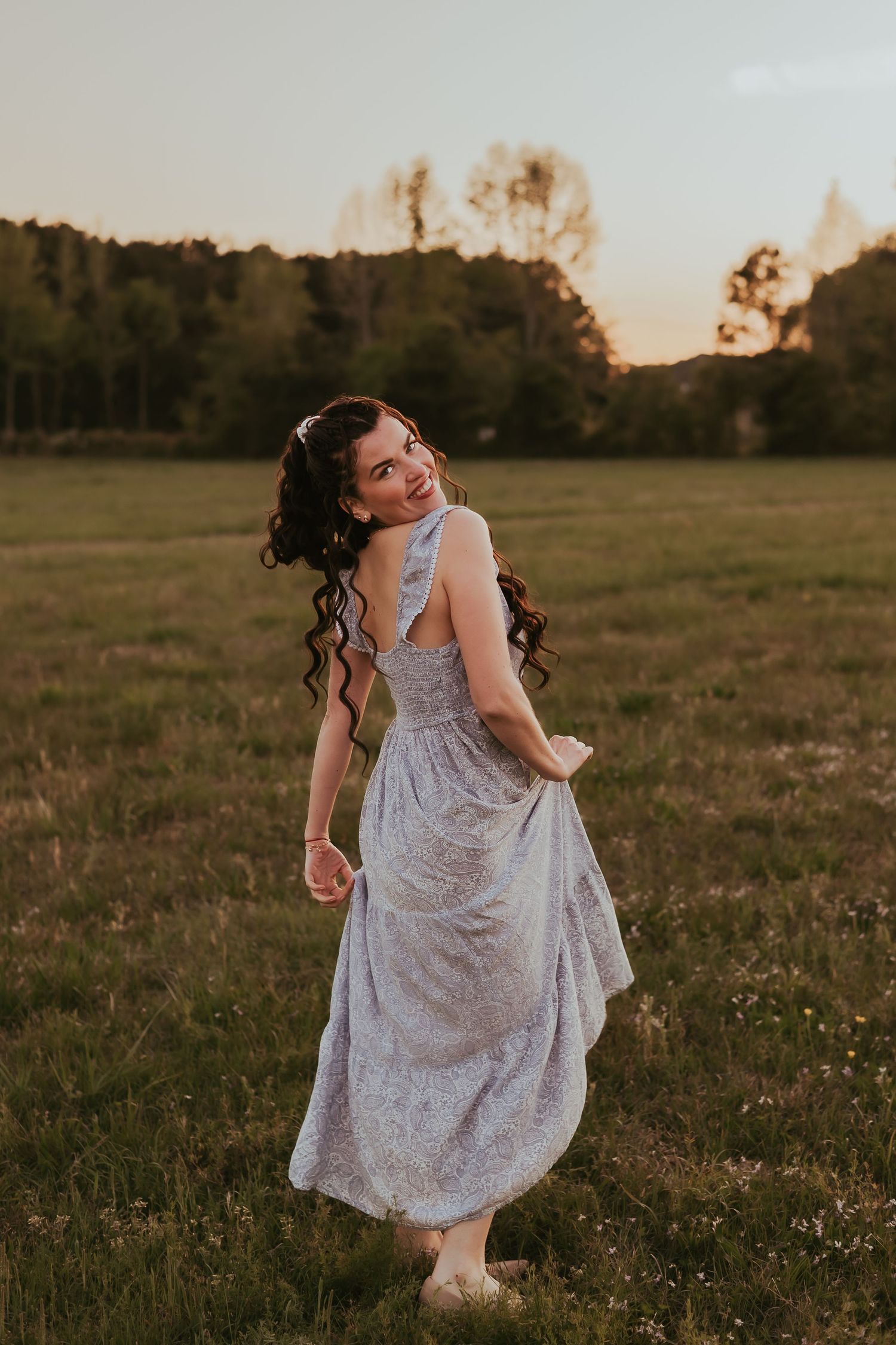 A woman in a light floral dress twirls playfully in a grassy field at sunset.