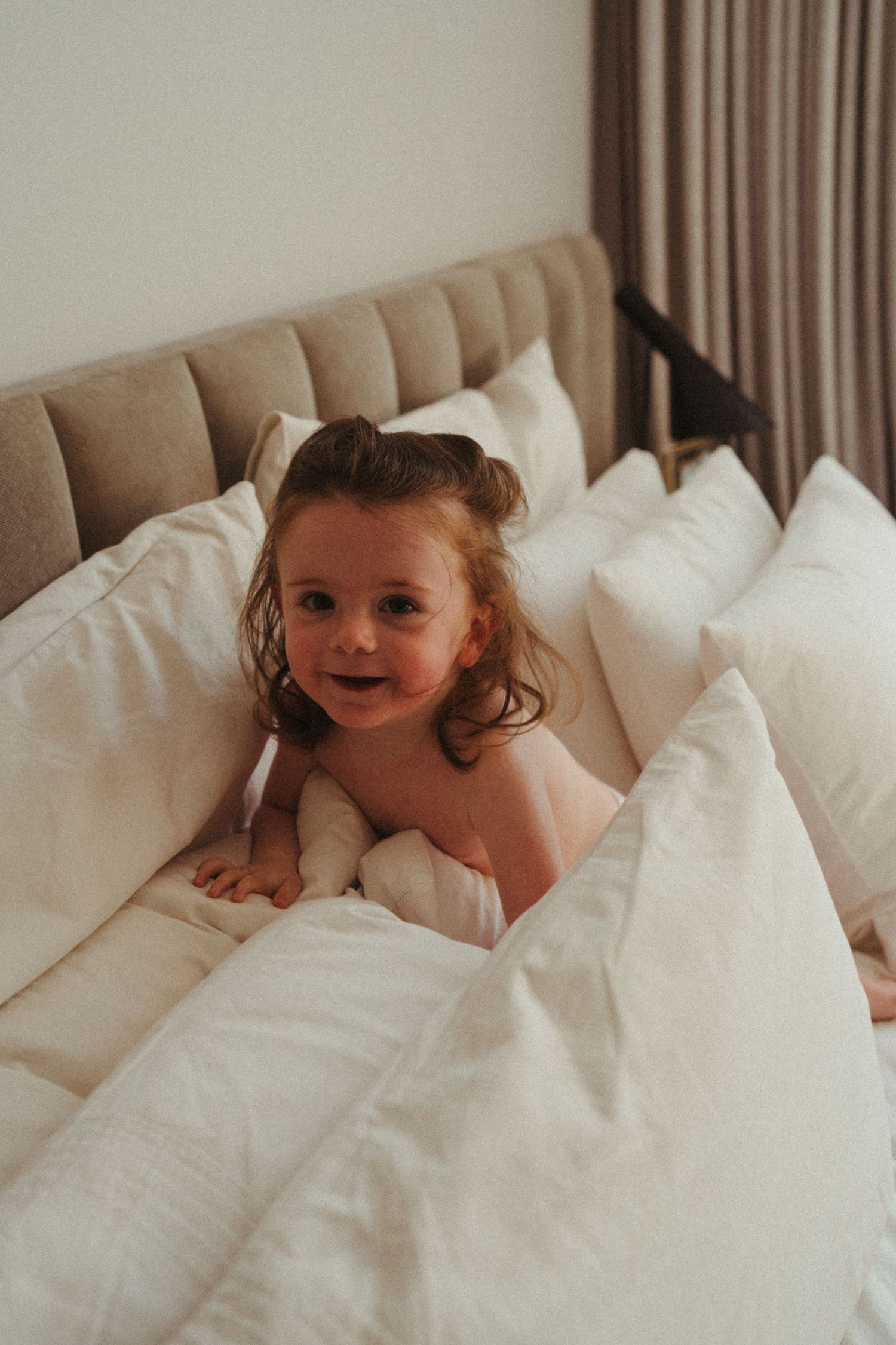 Child playing peek-a-boo under white bedding in a cozy bedroom setting.