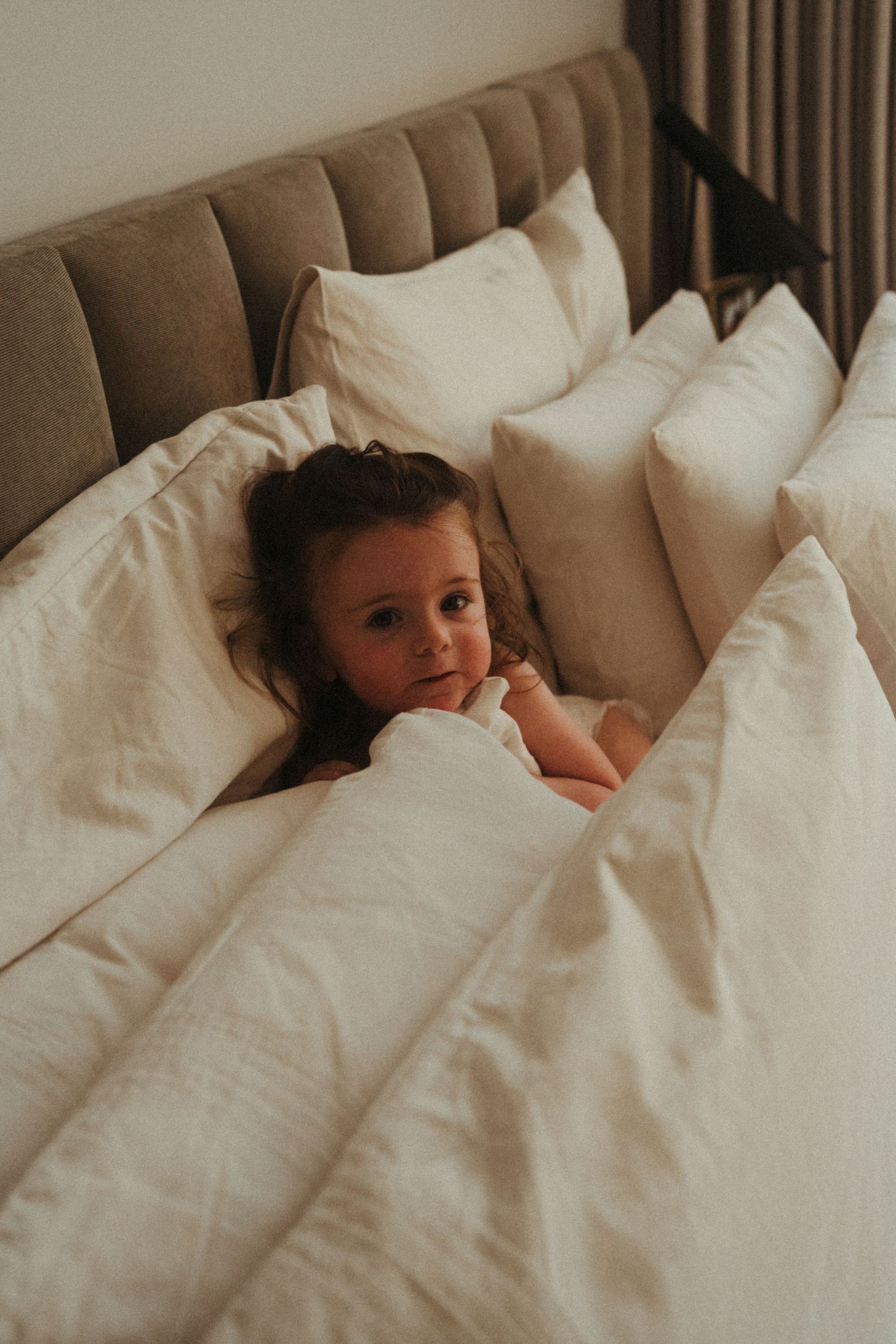Child peeking out from under white bedding and pillows in a cozy bedroom setting.