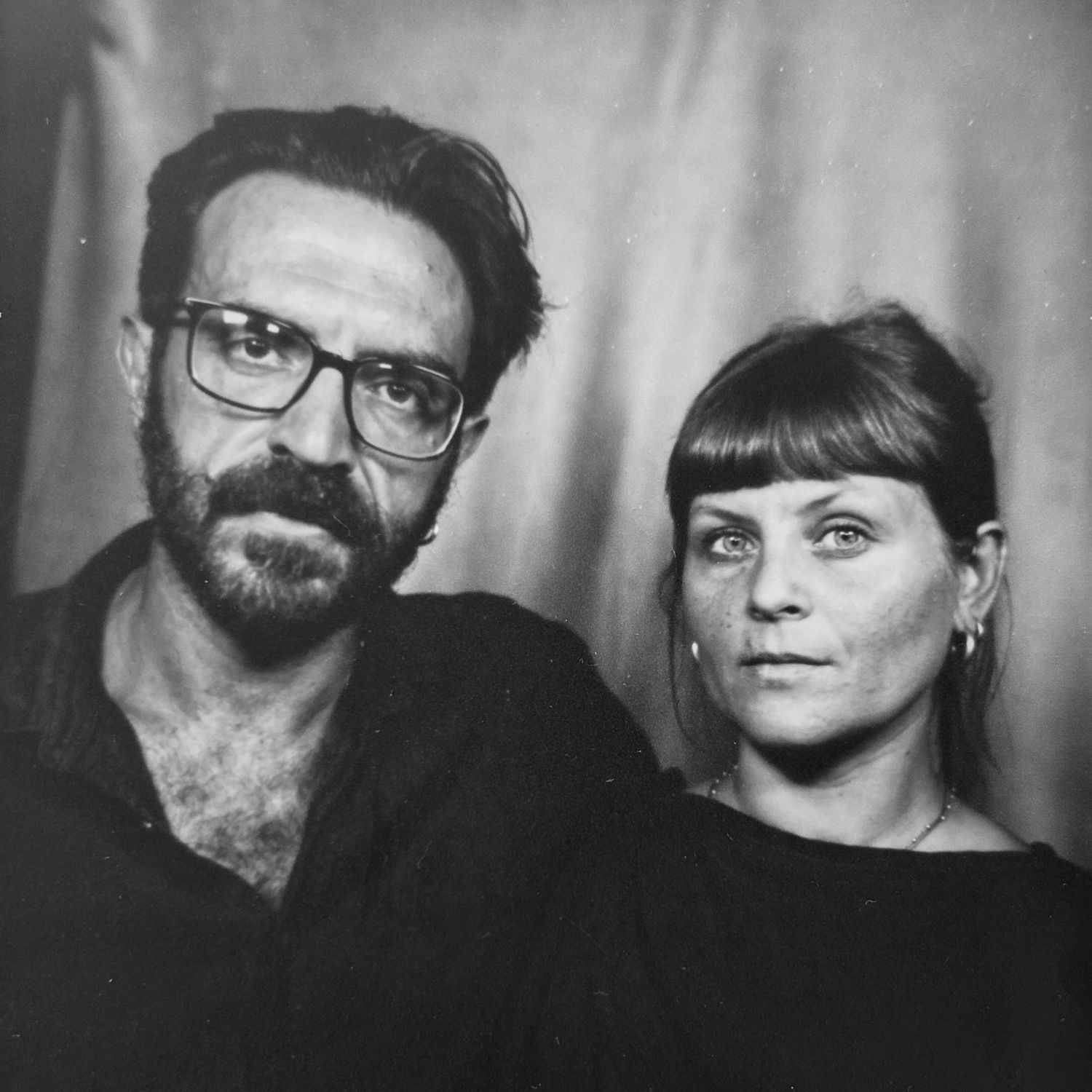 Black and white photo booth portrait of a bearded man with glasses and a woman with bangs, both wearing dark clothing.
