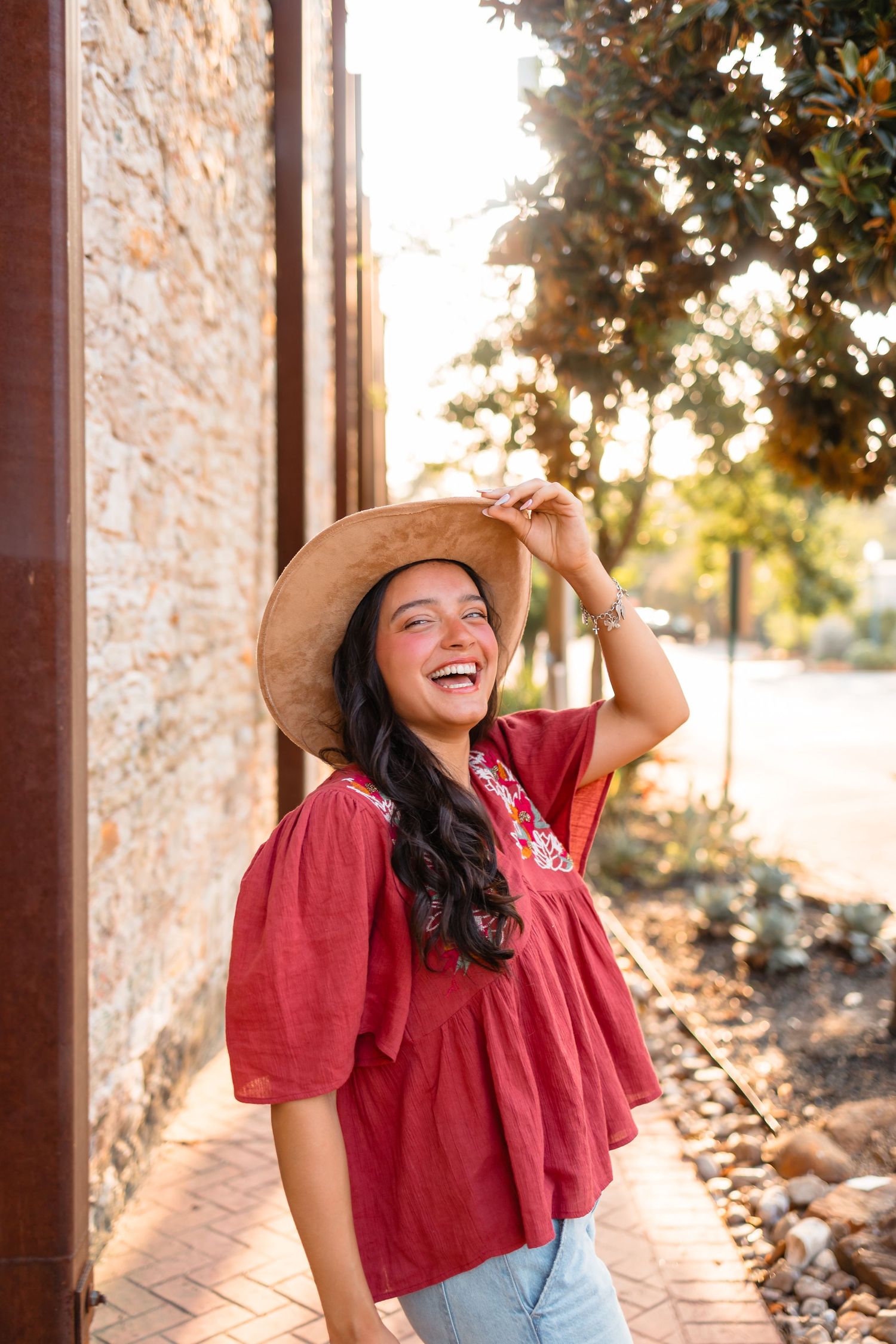 Laughing young woman in a red blouse and straw hat posing beside a stone wall outdoors.