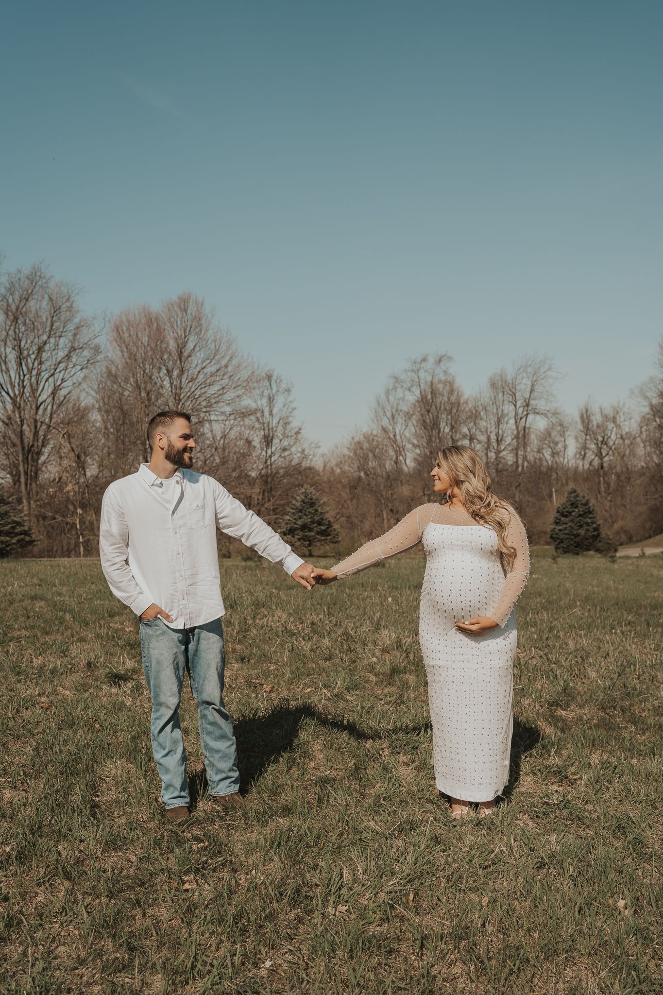 An expectant couple holds hands in a sunny field with Michigan fall foliage in the background.