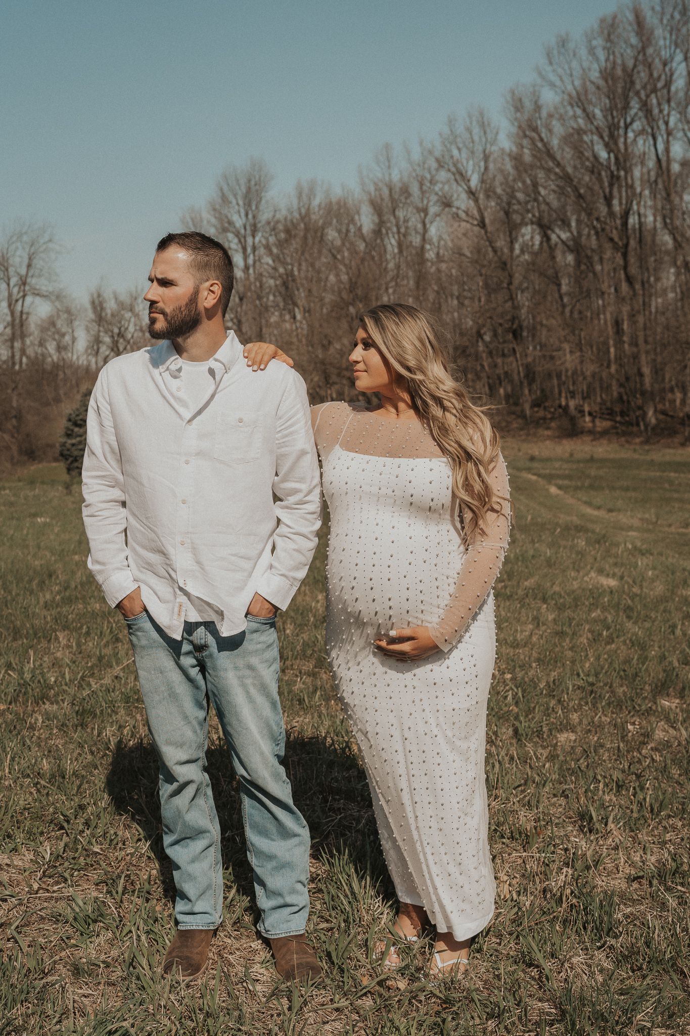 An outdoor maternity photo of a couple in casual white outfits standing in a grassy field.