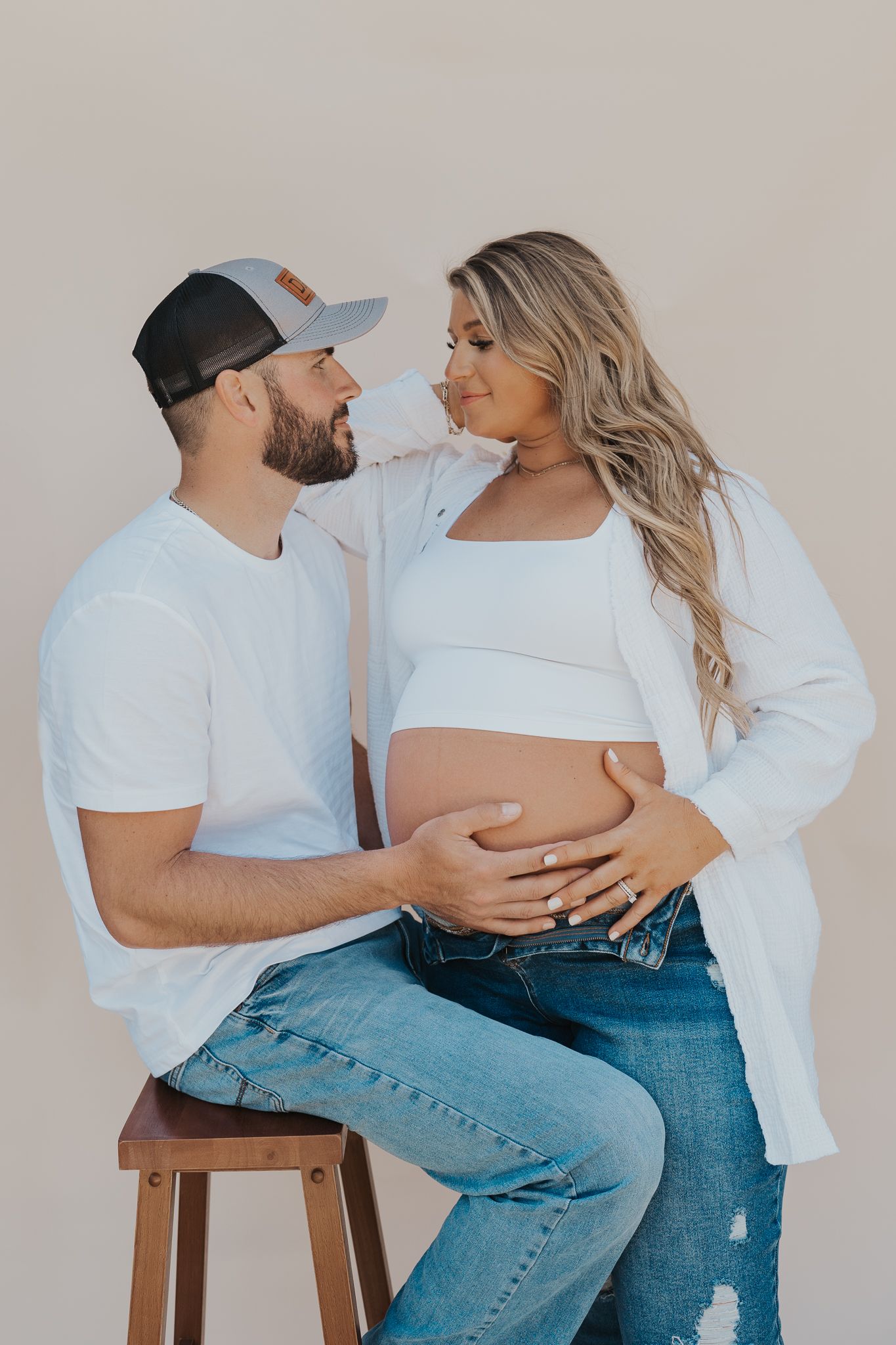An intimate studio maternity photo with a couple in white clothing and denim against a neutral background.
