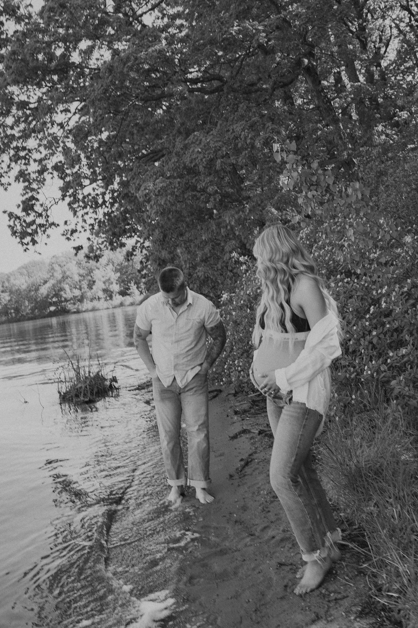 A black and white maternity photo of a couple by a lake shore.