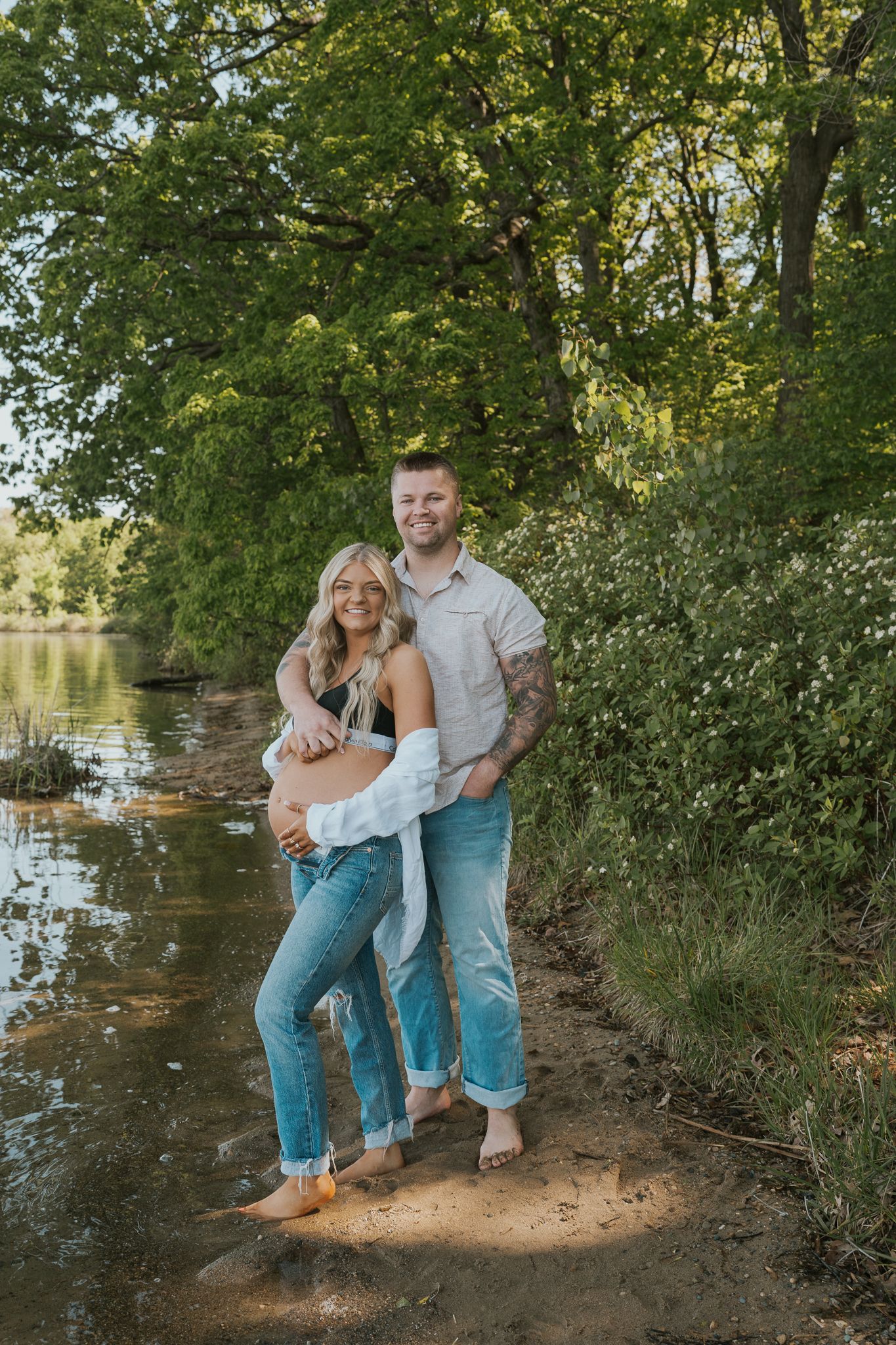 A casual maternity photo of a couple posing by a lake with green trees in the background taken by a Michigan based photographer.