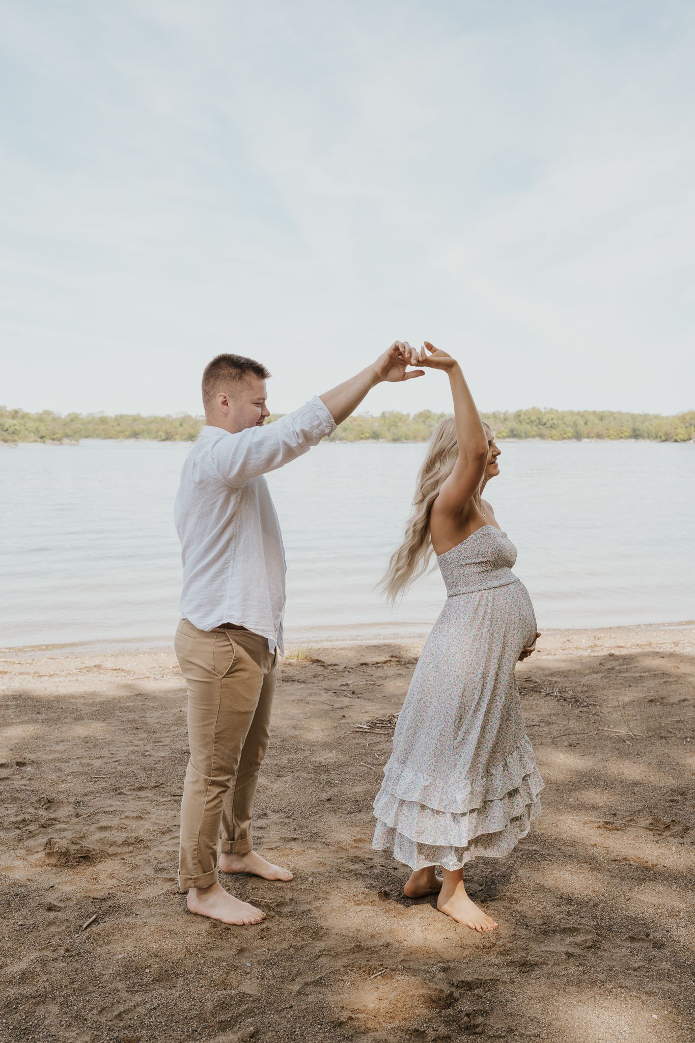 Playful beach maternity photos with couple making heart shapes against ocean backdrop.