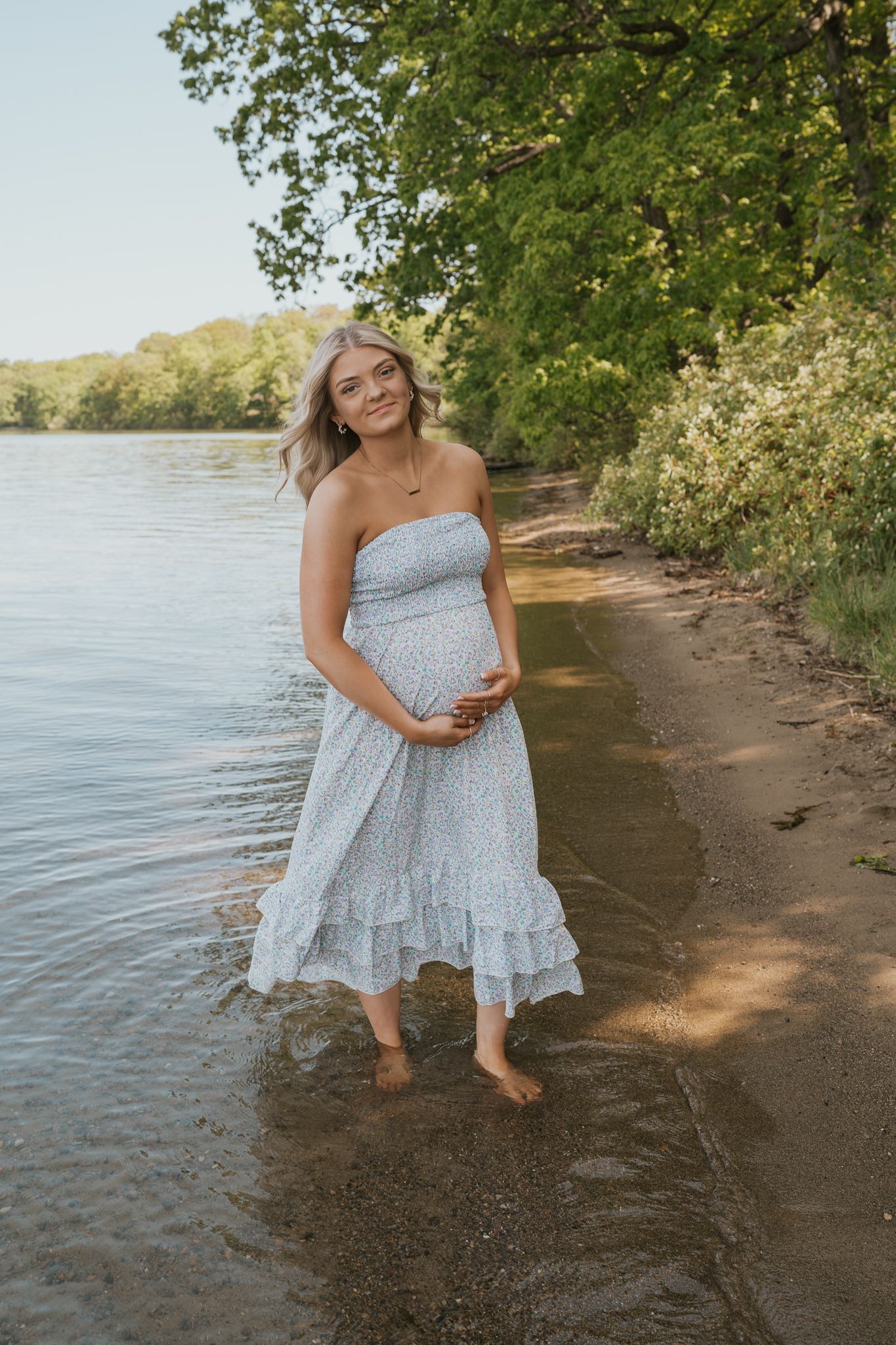 Expectant mother in flowy dress poses by Indiana waterfront during golden hour.