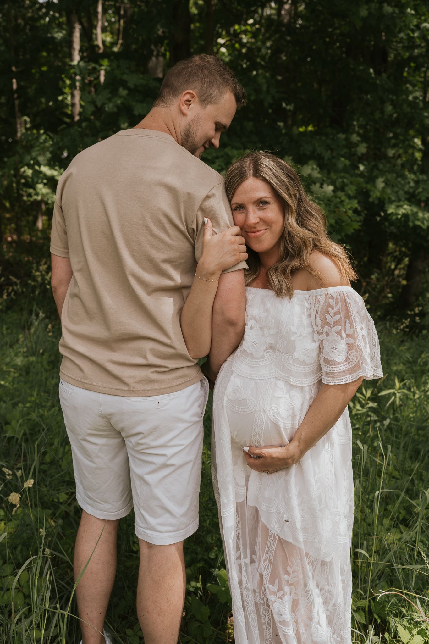 Romantic expecting couple in white and beige outfits embrace in a lush green outdoor setting.
