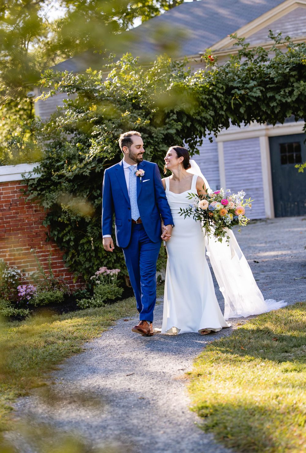Couple walking along a garden path outside a blue building with brick accents.