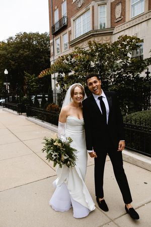 A couple poses on a city sidewalk in elegant wedding attire.